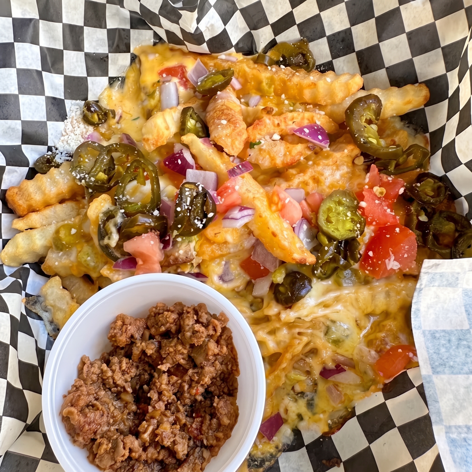 Loaded cheese fries topped with jalapeños, tomatoes, red onions, and a side of crispy seasoned meat in a bowl.