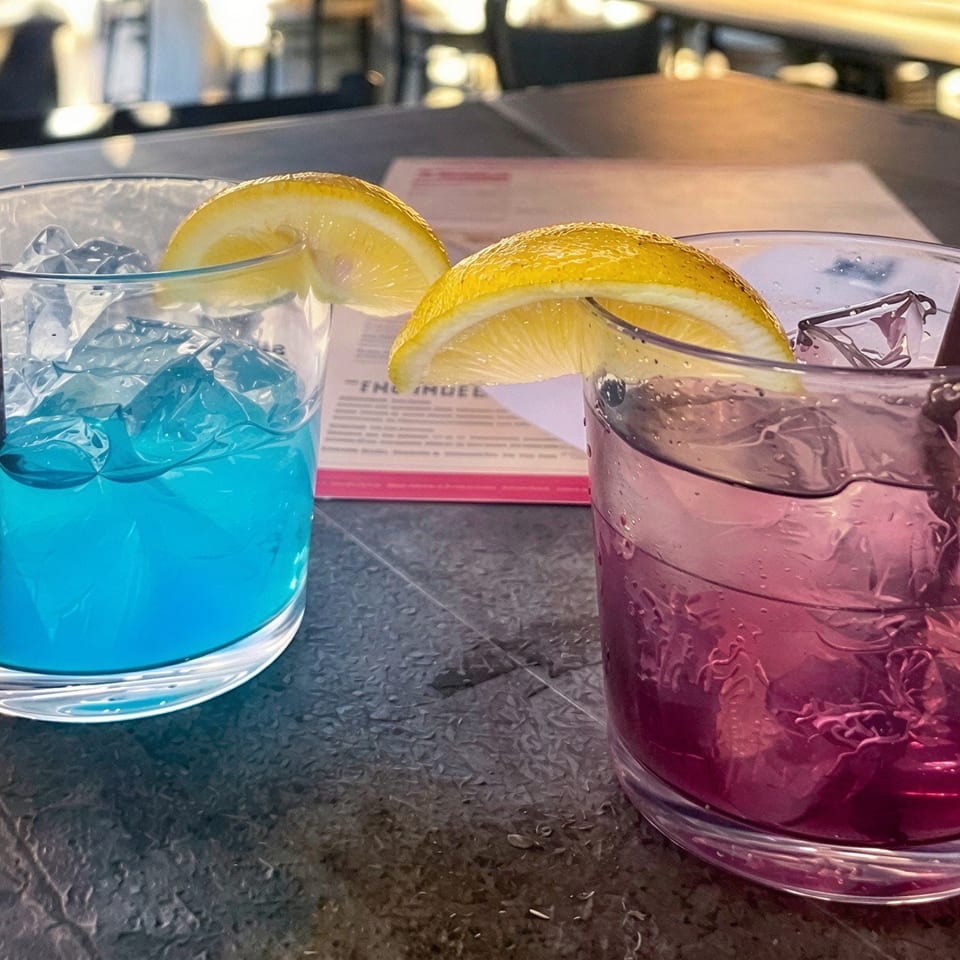 Two colorful iced cocktails, blue and purple, garnished with lemon wedges and served with straws at a restaurant table.