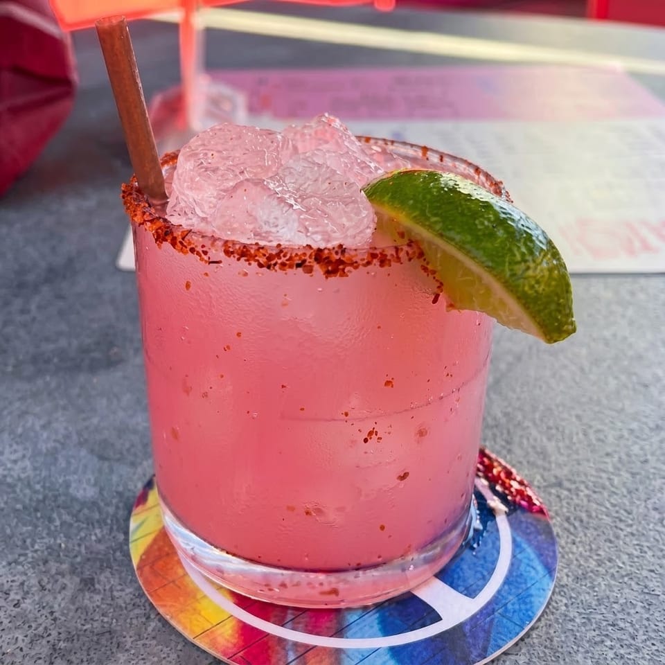 Pink iced cocktail with a chili-salt rim, lime wedge, and wooden straw on a colorful coaster.