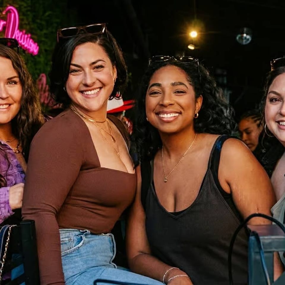 Four smiling women pose together outside a lively restaurant beneath a pink neon sign.