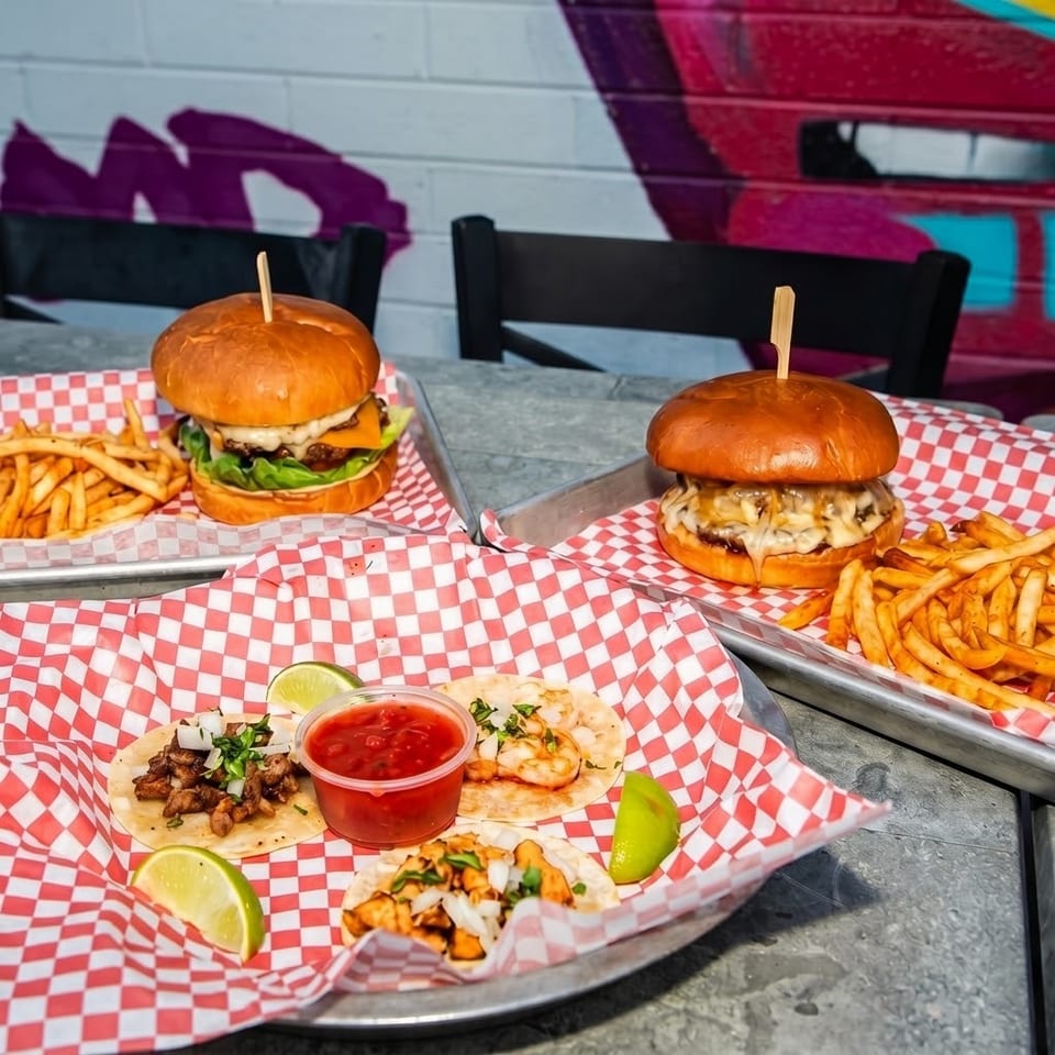 Two cheeseburgers with fries and tacos served on red-and-white checkered trays at an outdoor restaurant table.