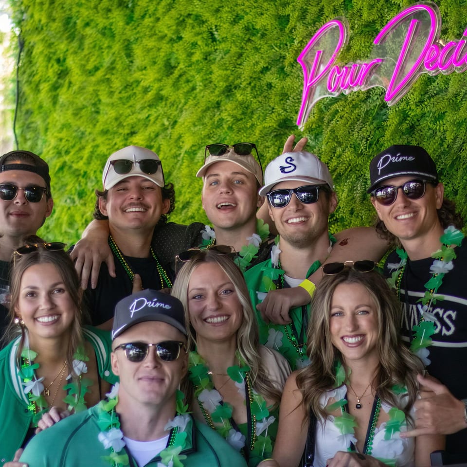 Group of friends wearing sunglasses, hats, and green leis pose beneath a neon “Prime Locations” sign against a leafy wall.