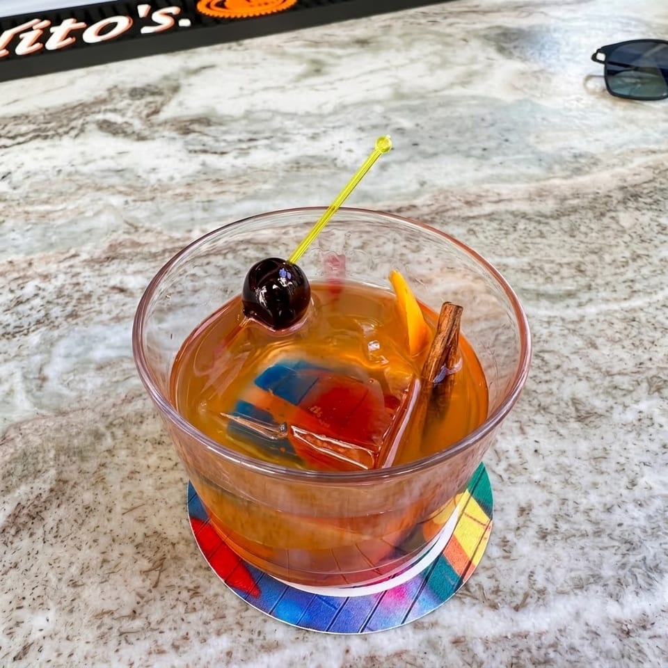 Amber cocktail garnished with a cherry, orange peel, cinnamon stick, and colorful ice cubes on a marble bar.