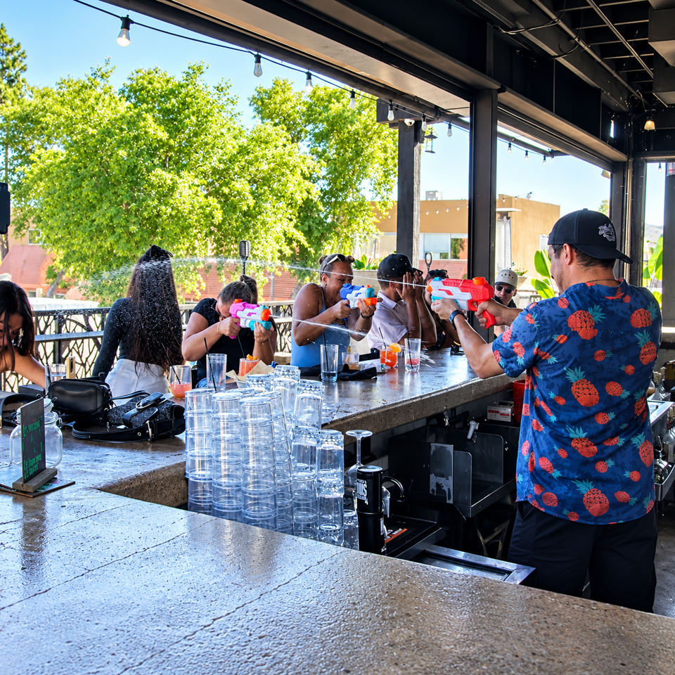 Restaurant guests playfully spray water guns across an open-air bar, surrounded by stacked glasses, drinks, string lights, and trees.