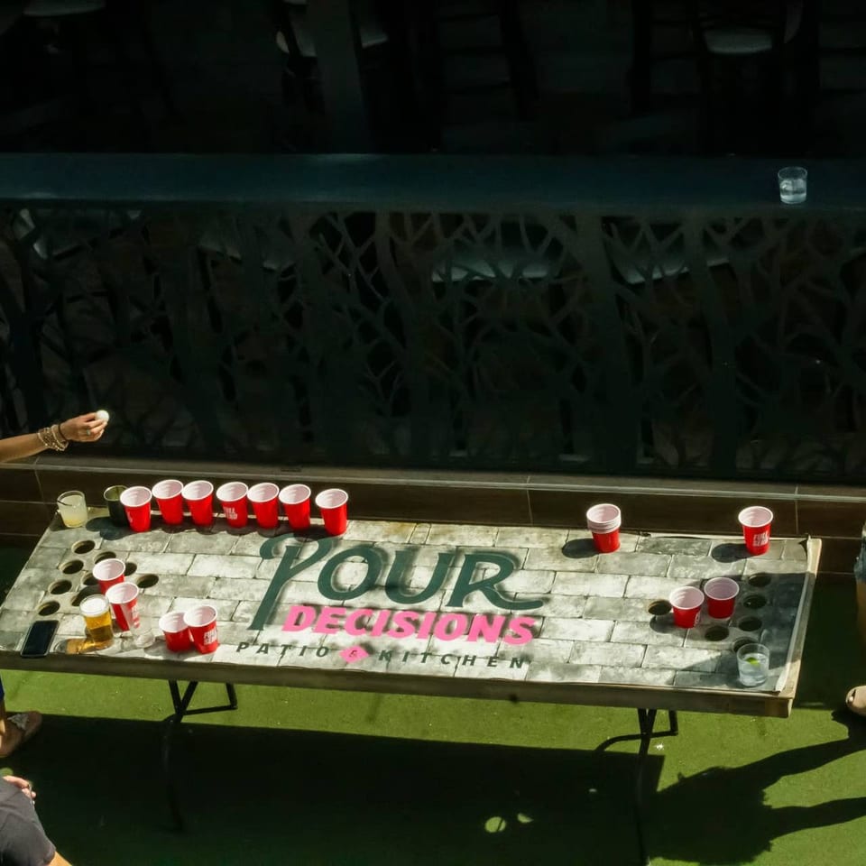 People play beer pong on a table branded “Four Decisions Patio Kitchen,” surrounded by red cups and drinks.