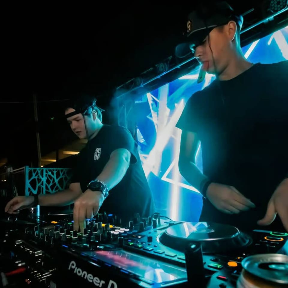 Two DJs perform at a club mixing console beneath blue geometric LED screens and stage lighting.