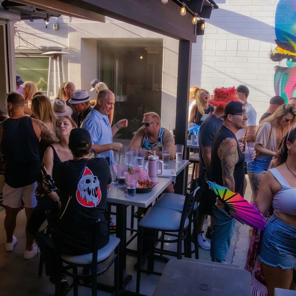 Busy restaurant patio filled with patrons, drinks, and snacks beneath string lights beside a colorful mural.