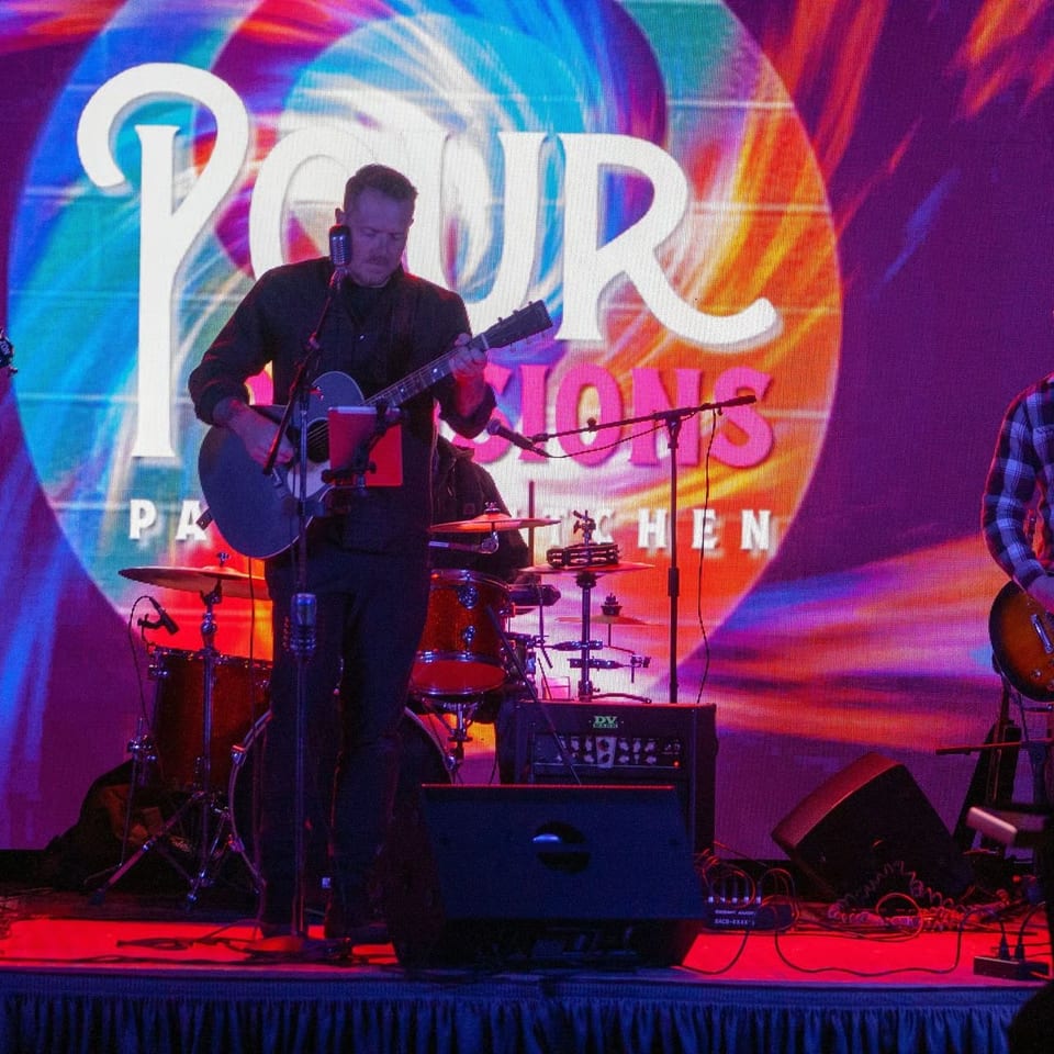 Three musicians perform onstage with guitars and drums under colorful lighting, backed by a large Power Hour Party logo.