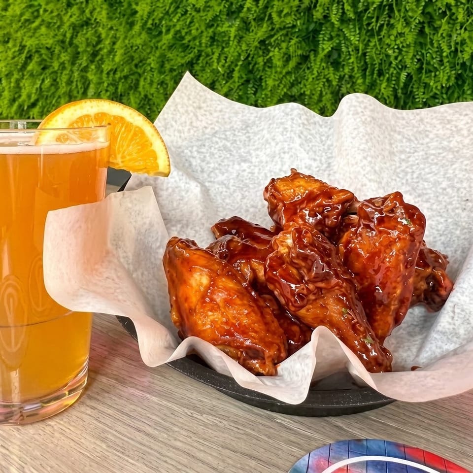 Glazed chicken wings in a paper-lined basket beside an orange-garnished drink on a wooden table.