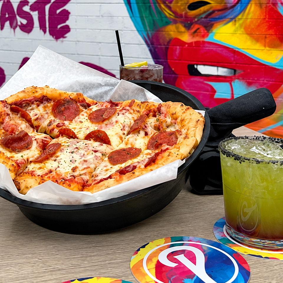 Pepperoni pizza in a black skillet with lime margarita, soda, and colorful graffiti wall behind.
