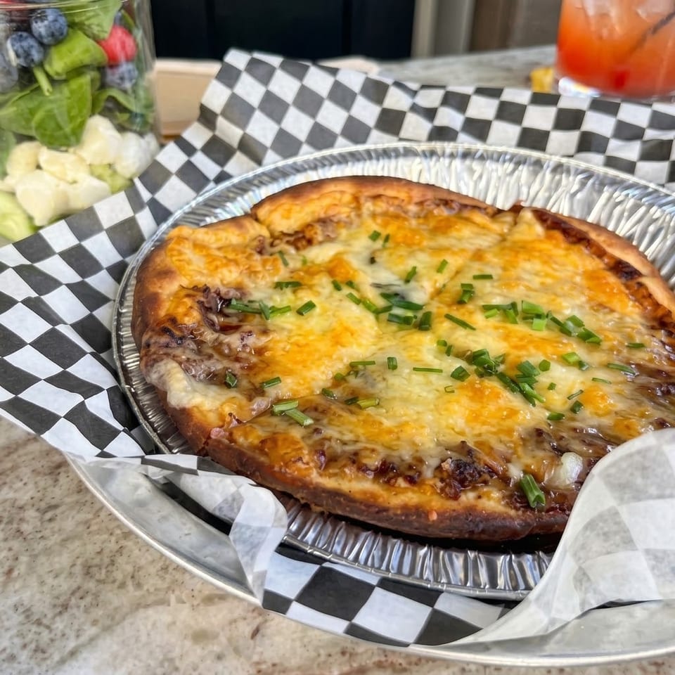 Cheesy baked pizza topped with chopped green onions, served in a foil tray lined with checkered paper.