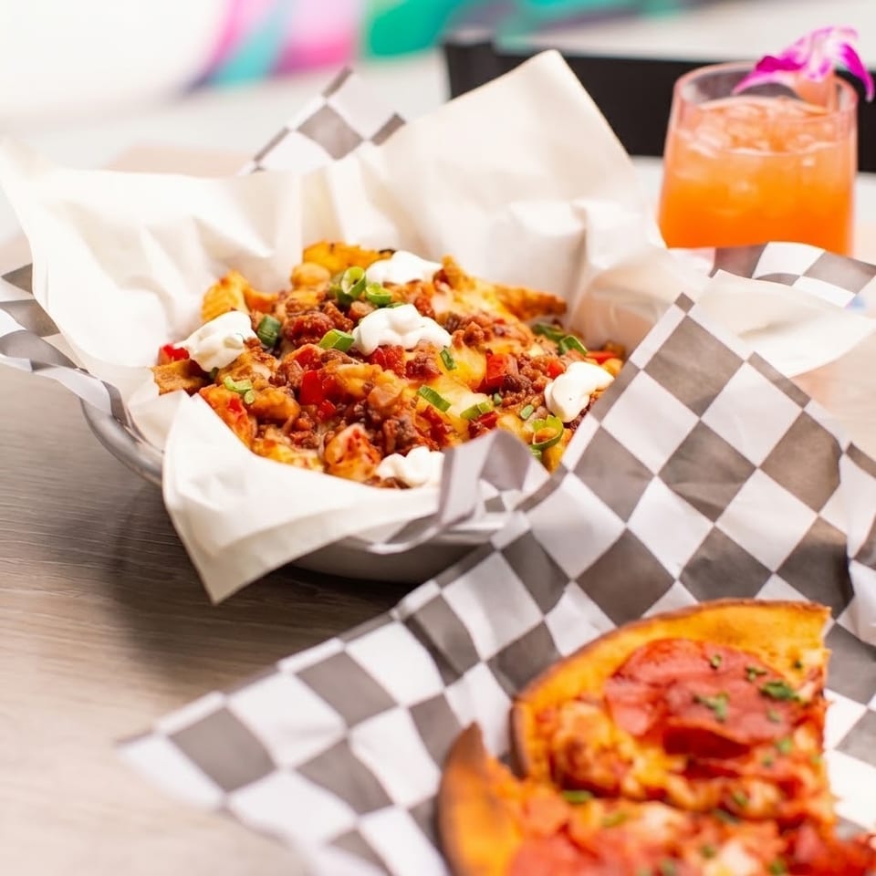 Loaded nachos with cheese, bacon, vegetables, and sour cream beside a pepperoni pizza and orange cocktail.